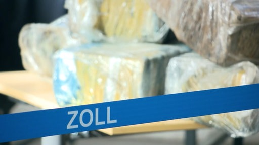 Im Vordergrund ist ein Banner mit der Aufschrift "Zoll" zu lesen. Im Hintergrund scheinbar Kokain-Päckchen.